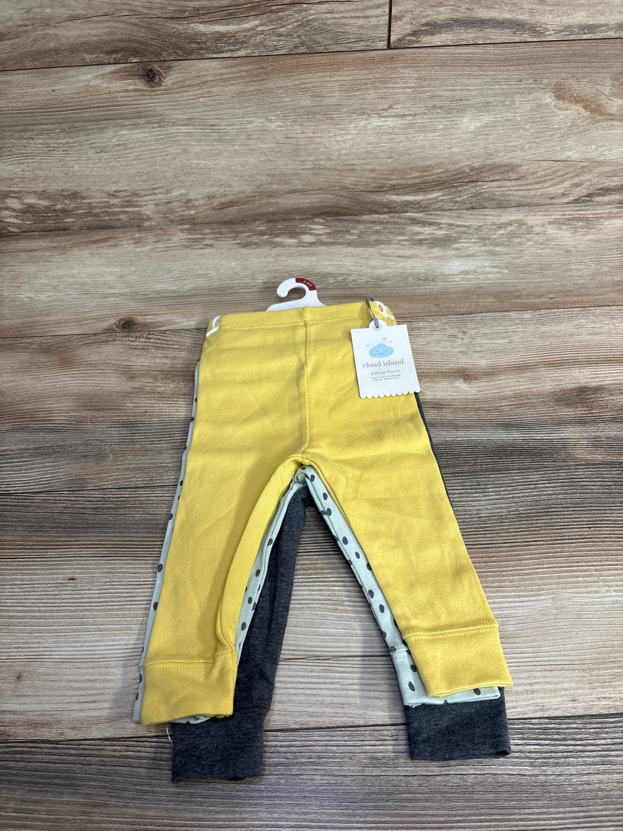 Three-pack of baby pants by Cloud Island in yellow, polka dot, and dark grey, laid on wood floor