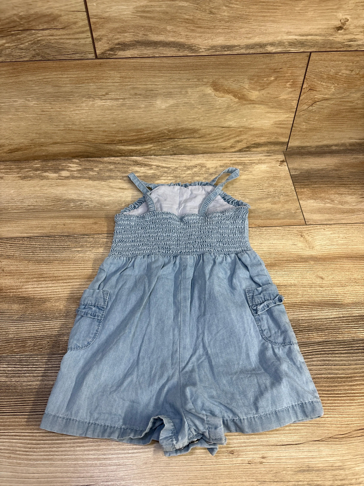 Front flat lay of blue chambray romper (18M) showing smocked bodice, thin straps, side pockets on wood floor