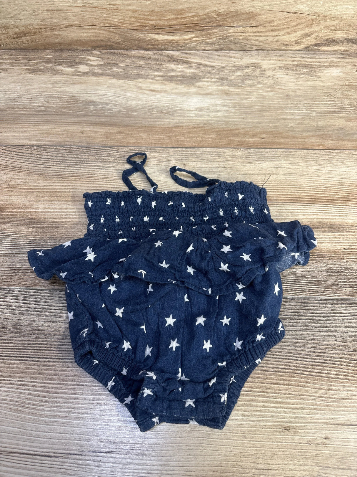 Top-down view of navy baby bubble romper with white stars, smocked ruffle top and shoulder ties on wood background