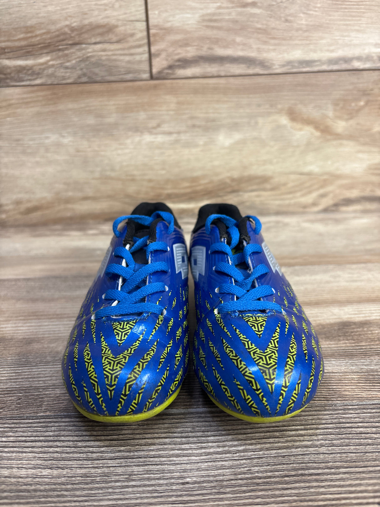 Front view of Lotto Forza Jr soccer cleats showing blue upper, yellow geometric pattern, and blue laces on wood floor