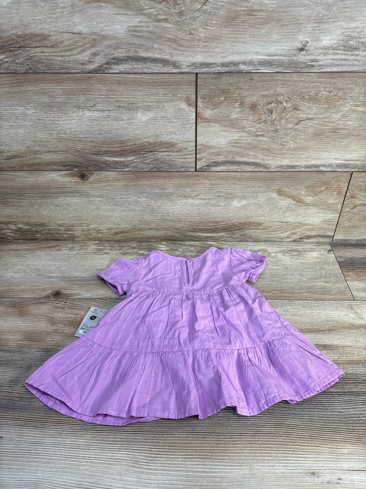 Flat lay of purple tiered baby dress showing short sleeves, gathered tiers and back keyhole on wood-plank background with tag visible