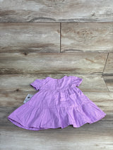 Flat lay of purple tiered baby dress showing short sleeves, gathered tiers and back keyhole on wood-plank background with tag visible