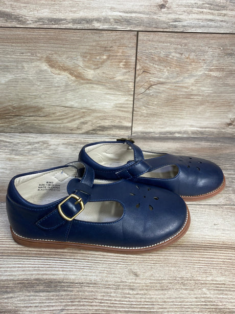 Side view of Felix & Flora navy T-strap Mary Jane flats showing gold buckle, floral toe cutouts, stitched tan sole, and visible size label on wood background