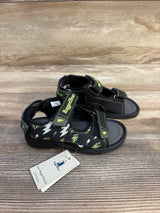 Three-quarter view of Rugged Bear toddler black open-toe sandals showcasing green trim, lightning-bolt print and Velcro straps on a wood surface