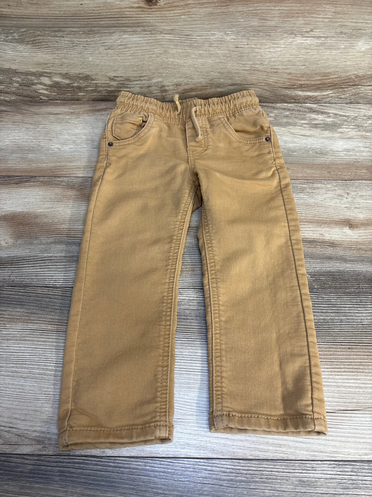 Tan kids pants with elastic waist, drawstring, and front pockets on wood floor