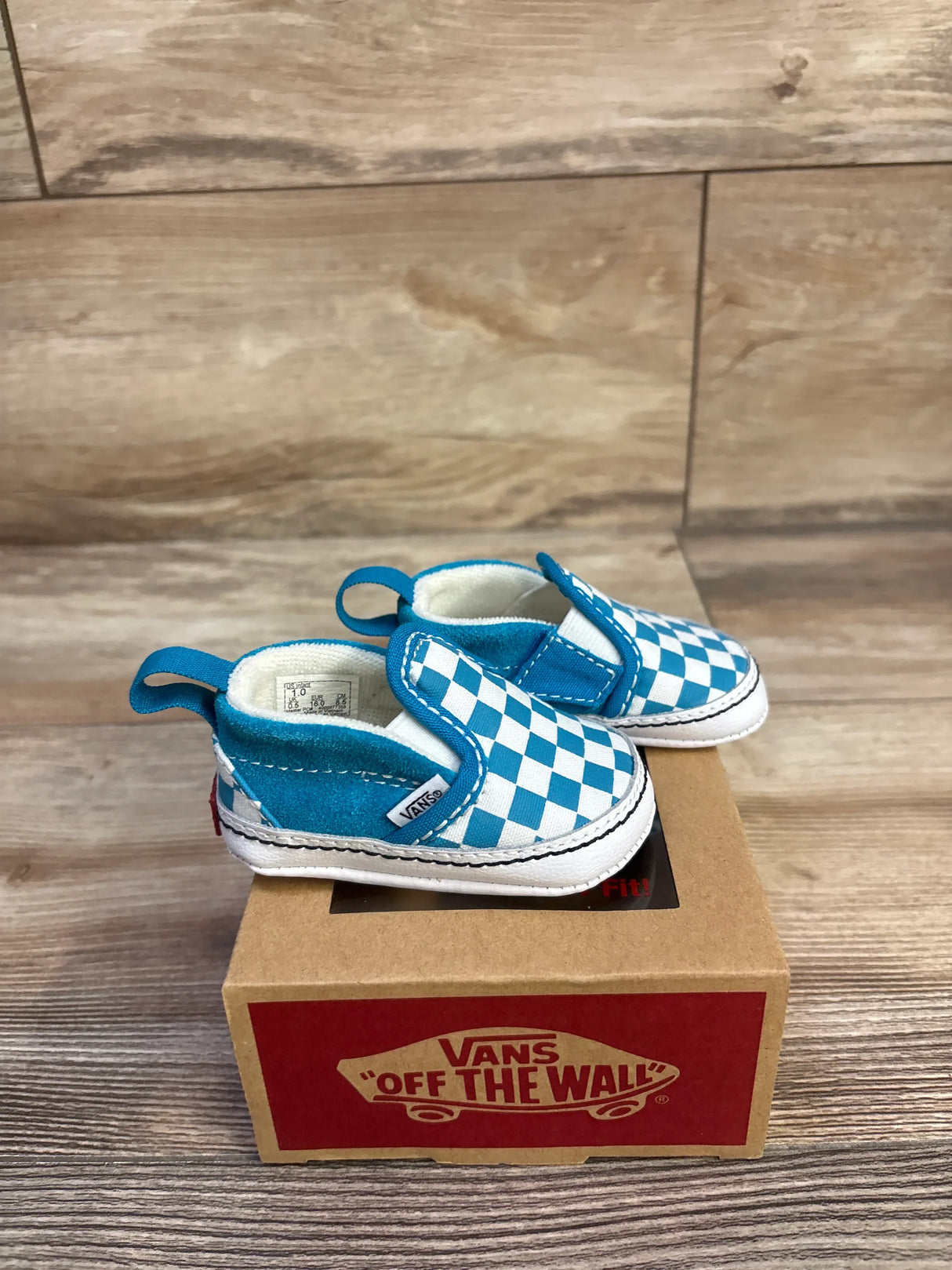 Angled front view of Vans baby slip-on crib shoes in blue checkerboard showing elastic side and pull tab on a Vans box