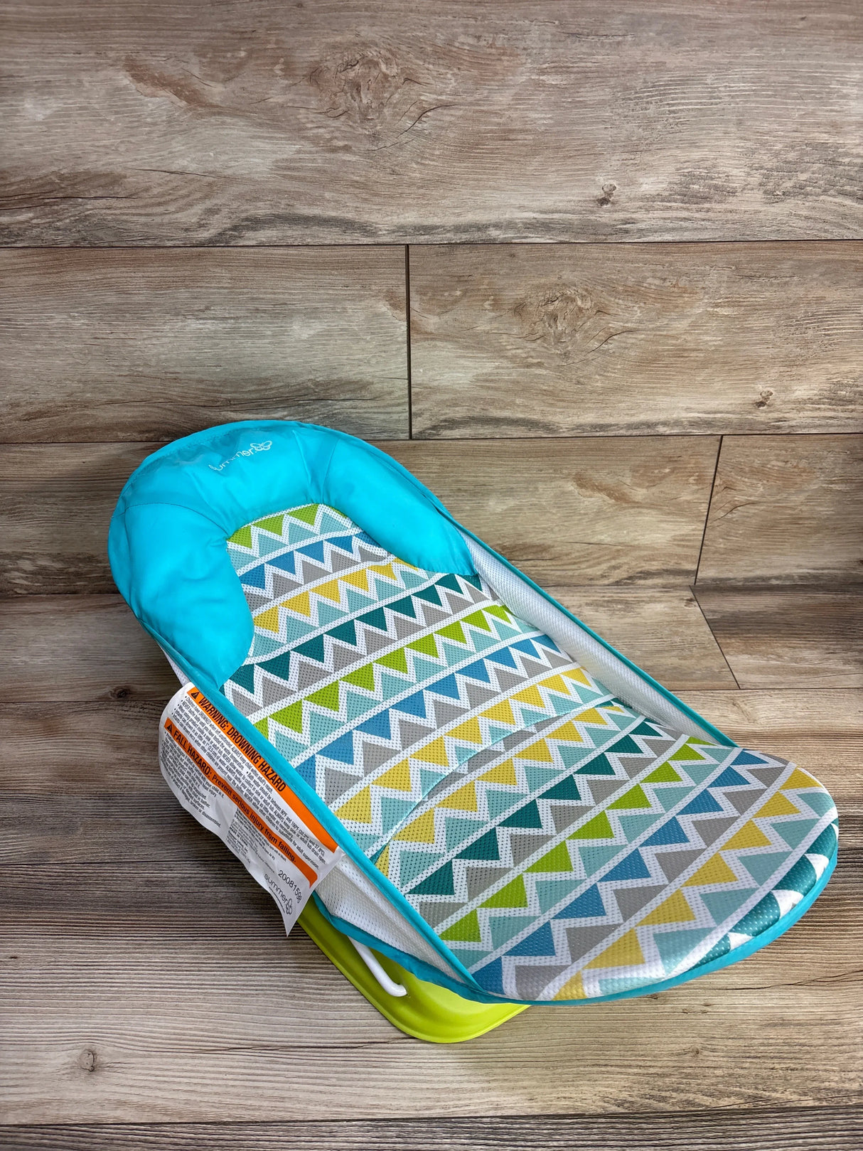 Angled view of Summer Deluxe baby bather in teal and green showing padded headrest, breathable mesh chevron seat and green plastic base on wood floor