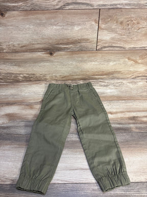 Front flat-lay of toddler olive green pull-on pants showing elastic waistband with drawstring, cuffed ankles, and side pockets on wood plank background