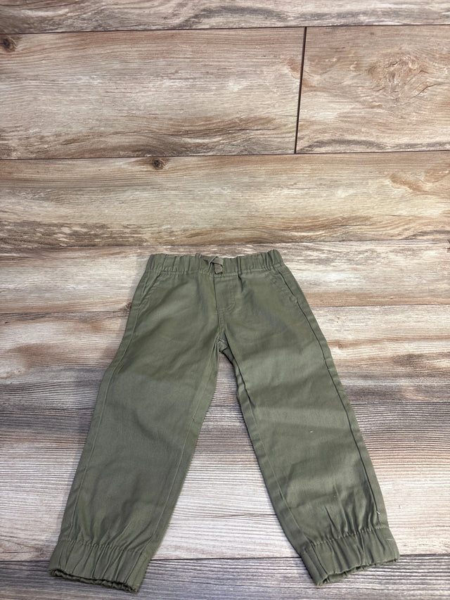 Front flat-lay of toddler olive green pull-on pants showing elastic waistband with drawstring, cuffed ankles, and side pockets on wood plank background
