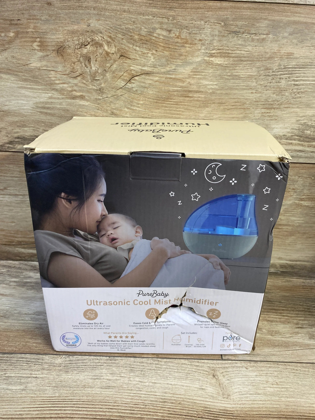 Front view of PureBaby Ultrasonic Cool Mist Humidifier boxed unit showing blue translucent tank and mother-and-baby image, dented cardboard box on wood background