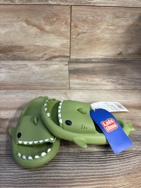Front three-quarter view of green Crayon Friends shark toddler slides showing open-toe shark-mouth teeth detail and blue product tag on wooden surface