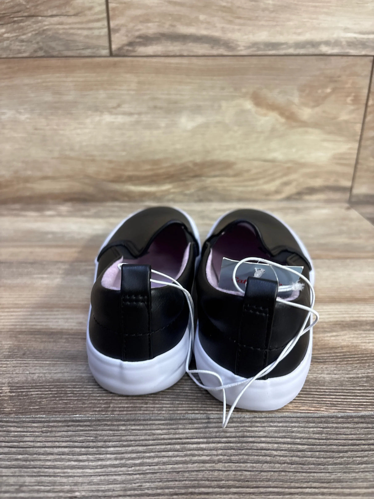 Rear view of Cat and Jack black toddler slip-on sneakers with white soles, heel pull tabs and pink lining on wood surface