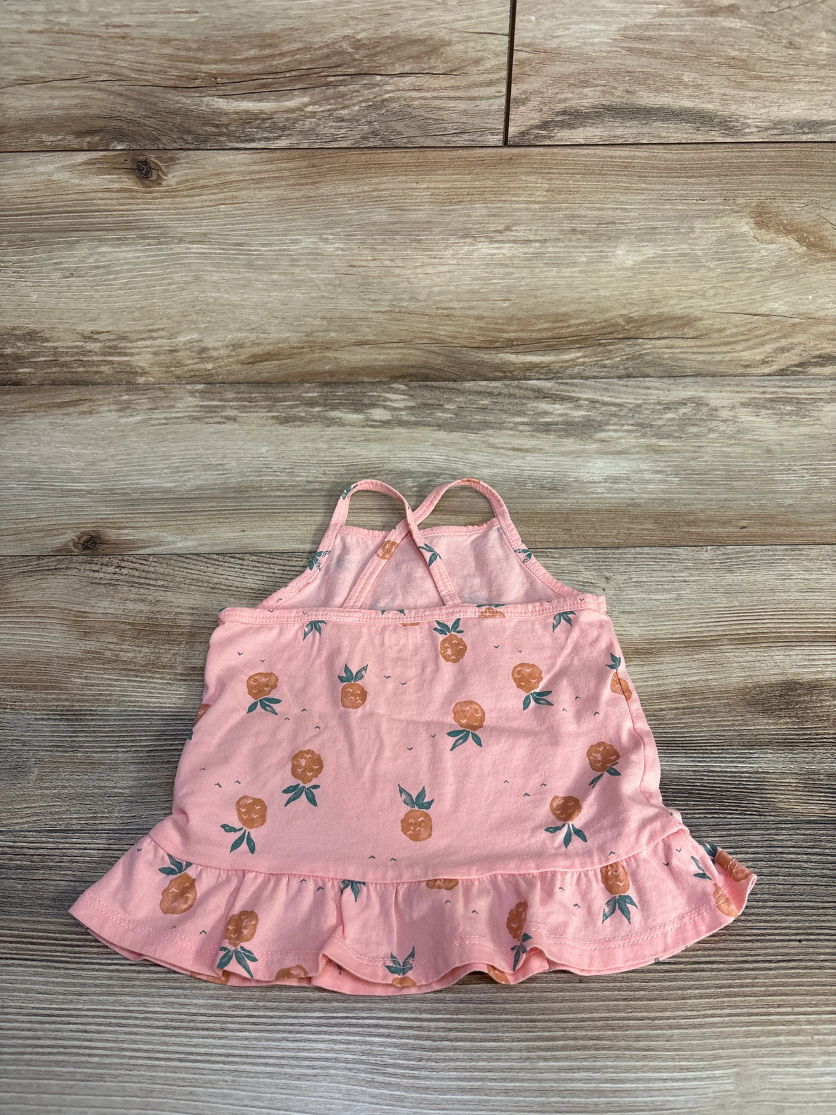 Flat lay of pink OshKosh 24-month tank top with pineapple print, ruffle hem, and crossed straps on light wood background
