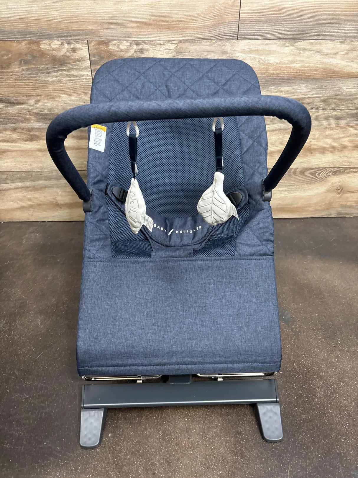 Front view of Baby Delight Alpine Deluxe bouncer in quilted indigo showcasing padded harness leaf toys and metal frame on wood plank floor