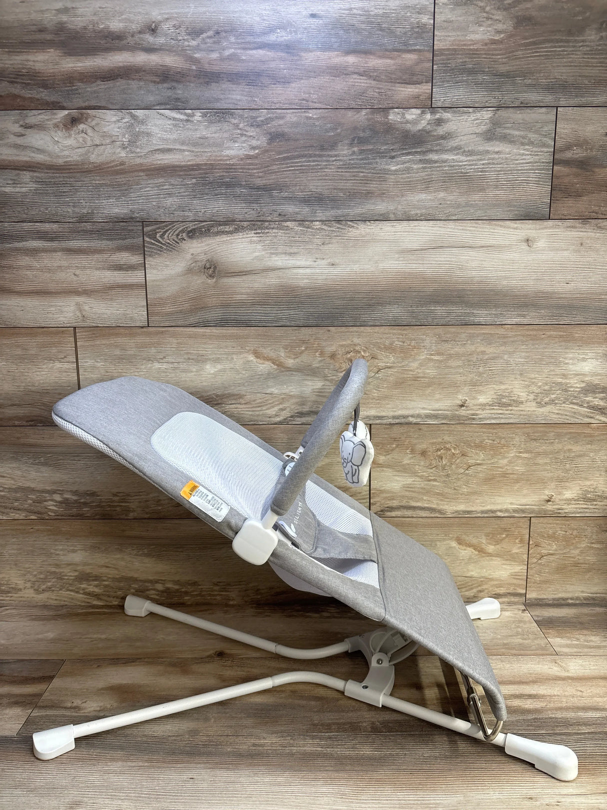 Side view of Baby Delight Highland baby bouncer showcasing pebble grey fabric, mesh center panel, toy bar with elephant plush, white metal frame on wood floor