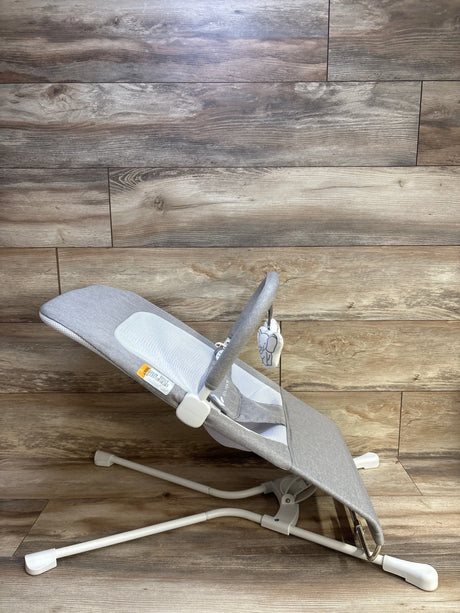 Side view of Baby Delight Highland baby bouncer showcasing pebble grey fabric, mesh center panel, toy bar with elephant plush, white metal frame on wood floor