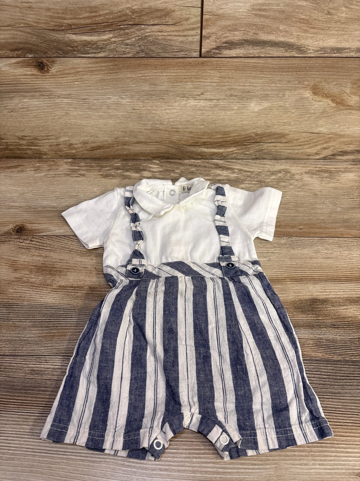 Flat-lay of white short-sleeve baby romper with navy striped suspender shorts, button straps and snap crotch on wood background