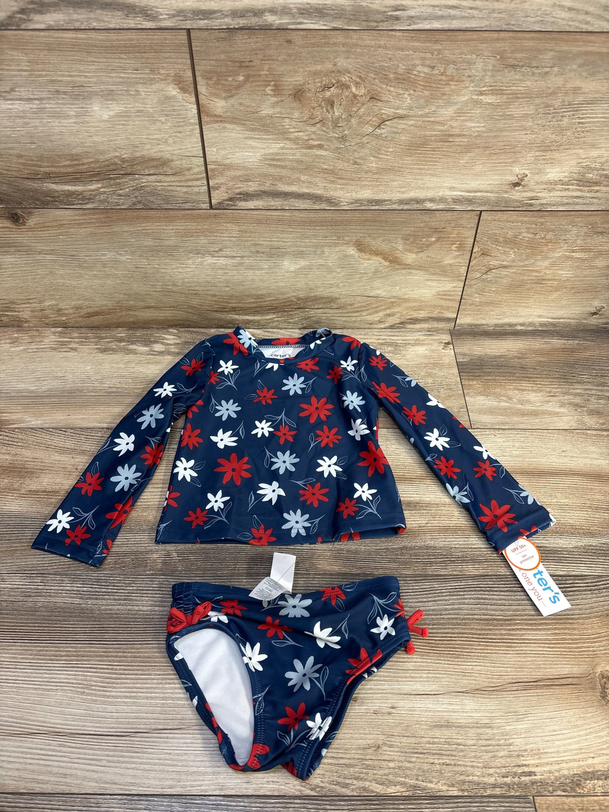 Flat lay of girls' navy 2-piece rash guard set showcasing red and white floral print on long-sleeve top and matching bikini bottoms with wood floor background