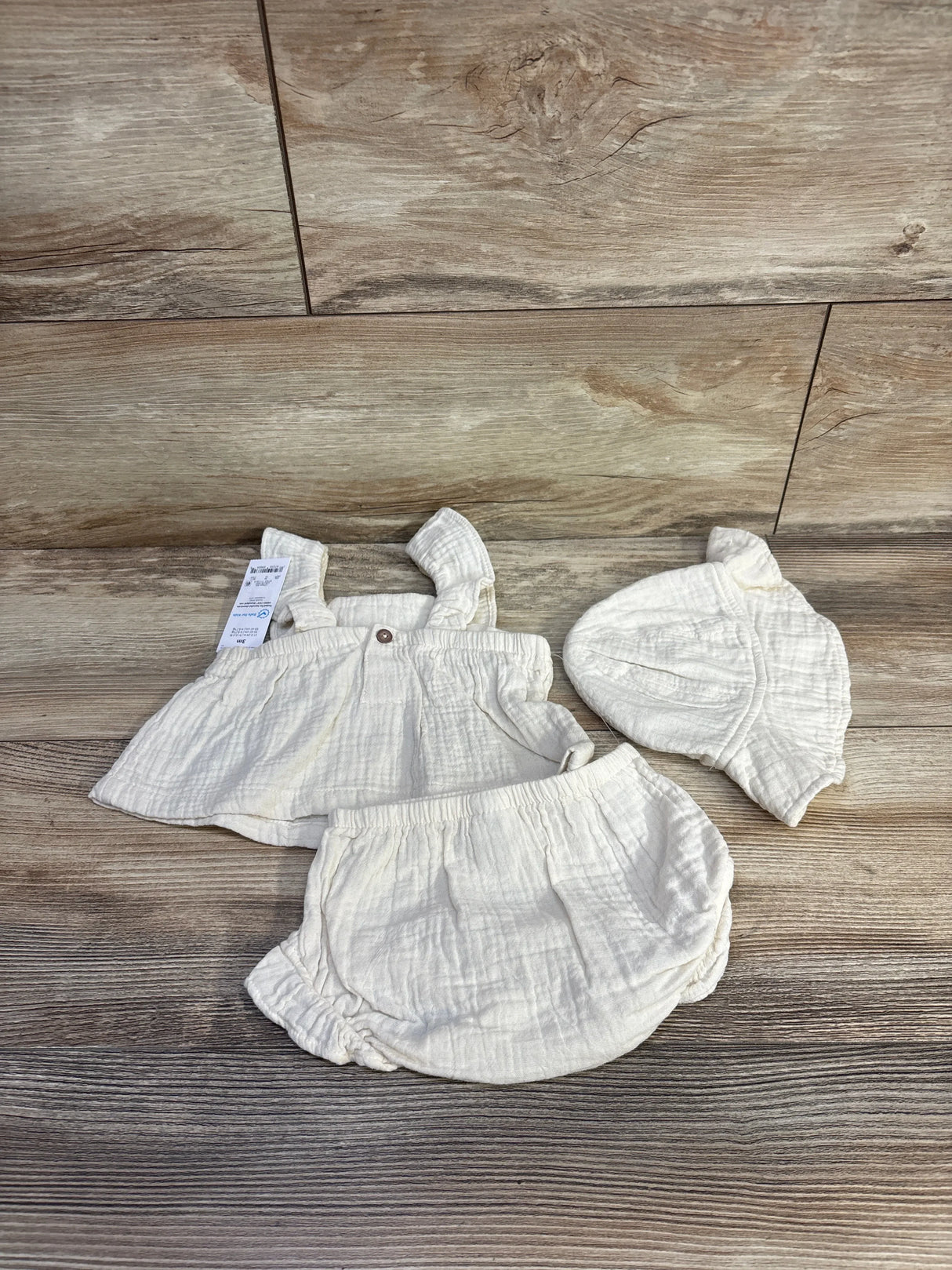 Flat lay of beige 3-piece baby set showing ruffled sleeveless top, elastic-waist bloomers, and matching hat on wood background