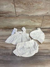 Flat lay of beige 3-piece baby set showing ruffled sleeveless top, elastic-waist bloomers, and matching hat on wood background