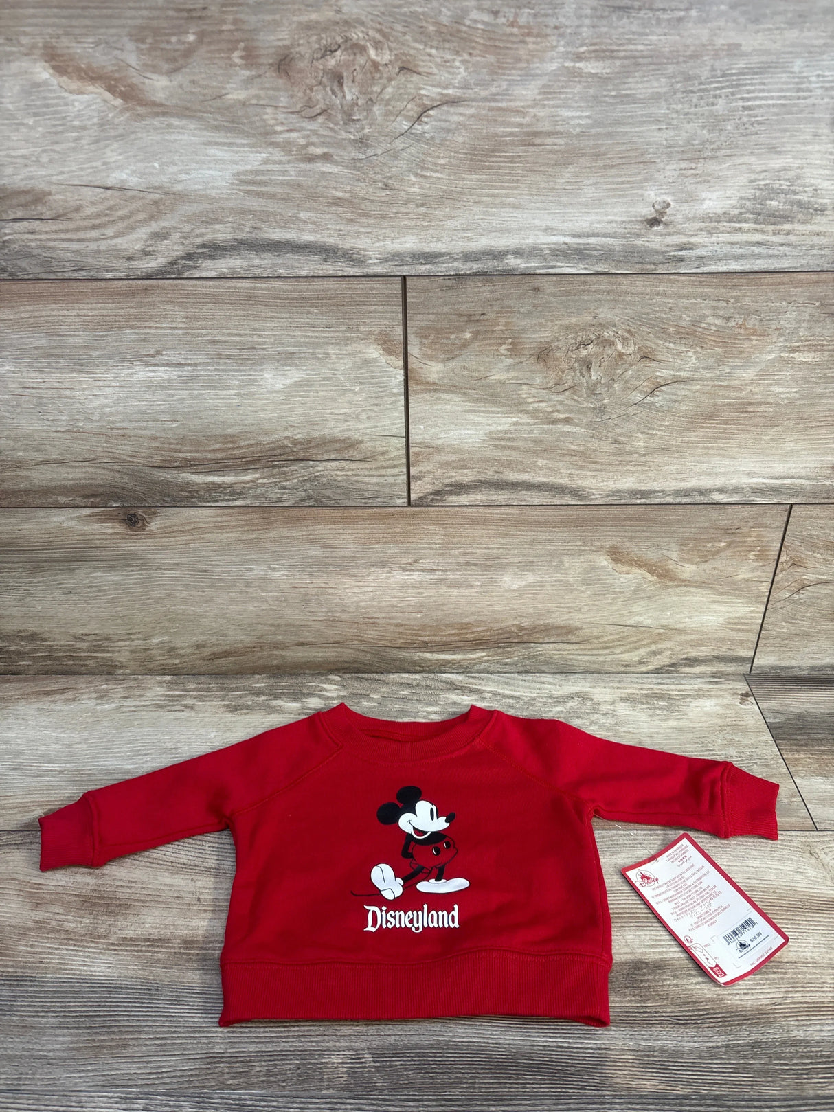 Front view of red baby sweatshirt with Mickey Mouse graphic and "Disneyland" text, ribbed cuffs and hem, size 3-6M tag on wood floor