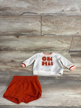Toddler two-piece outfit with white long-sleeve top featuring orange "OH Hi!" text and orange shorts on wood floor