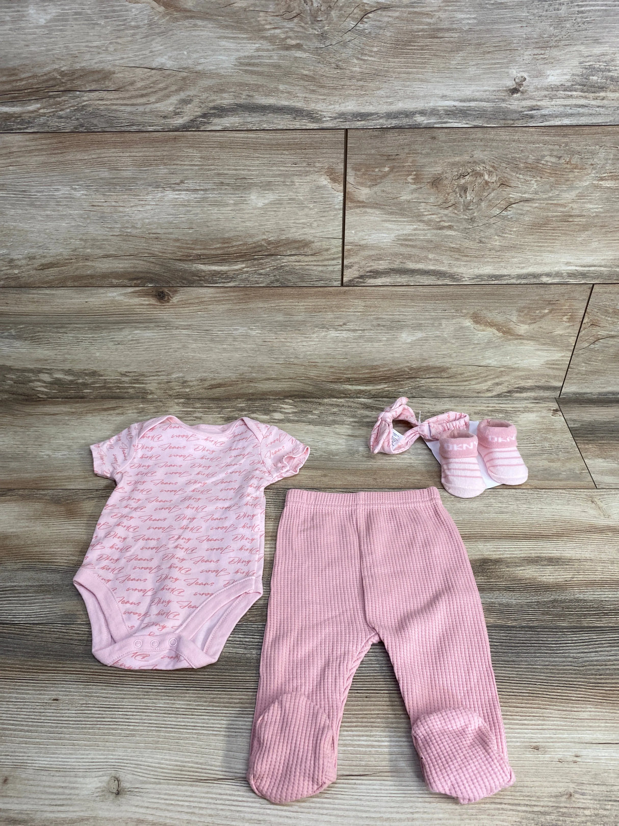 Pink baby outfit set with a logo print onesie, footed pants, matching headband, and socks on wood floor