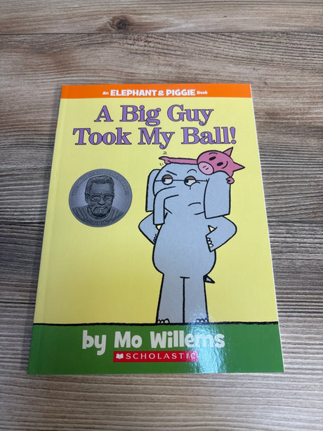 Front view of A Big Guy Took My Ball Elephant and Piggie book by Mo Willems with gray elephant carrying pink pig on yellow cover on wood plank floor