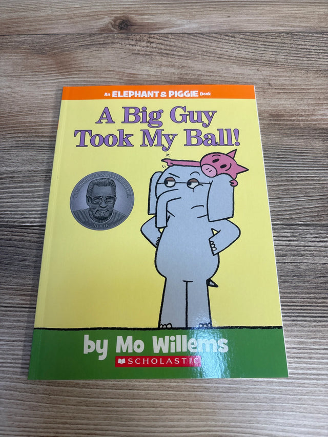Front view of A Big Guy Took My Ball Elephant and Piggie book by Mo Willems with gray elephant carrying pink pig on yellow cover on wood plank floor