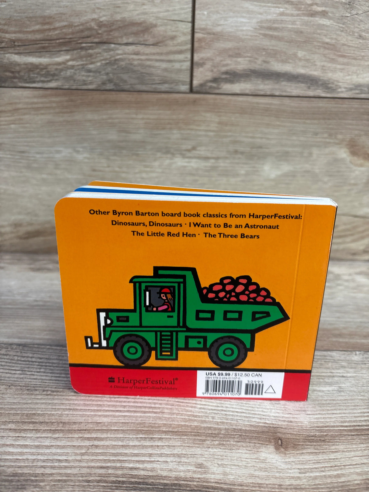 Back view of Machines At Work board book showing green dump truck illustration on orange background on wood floor