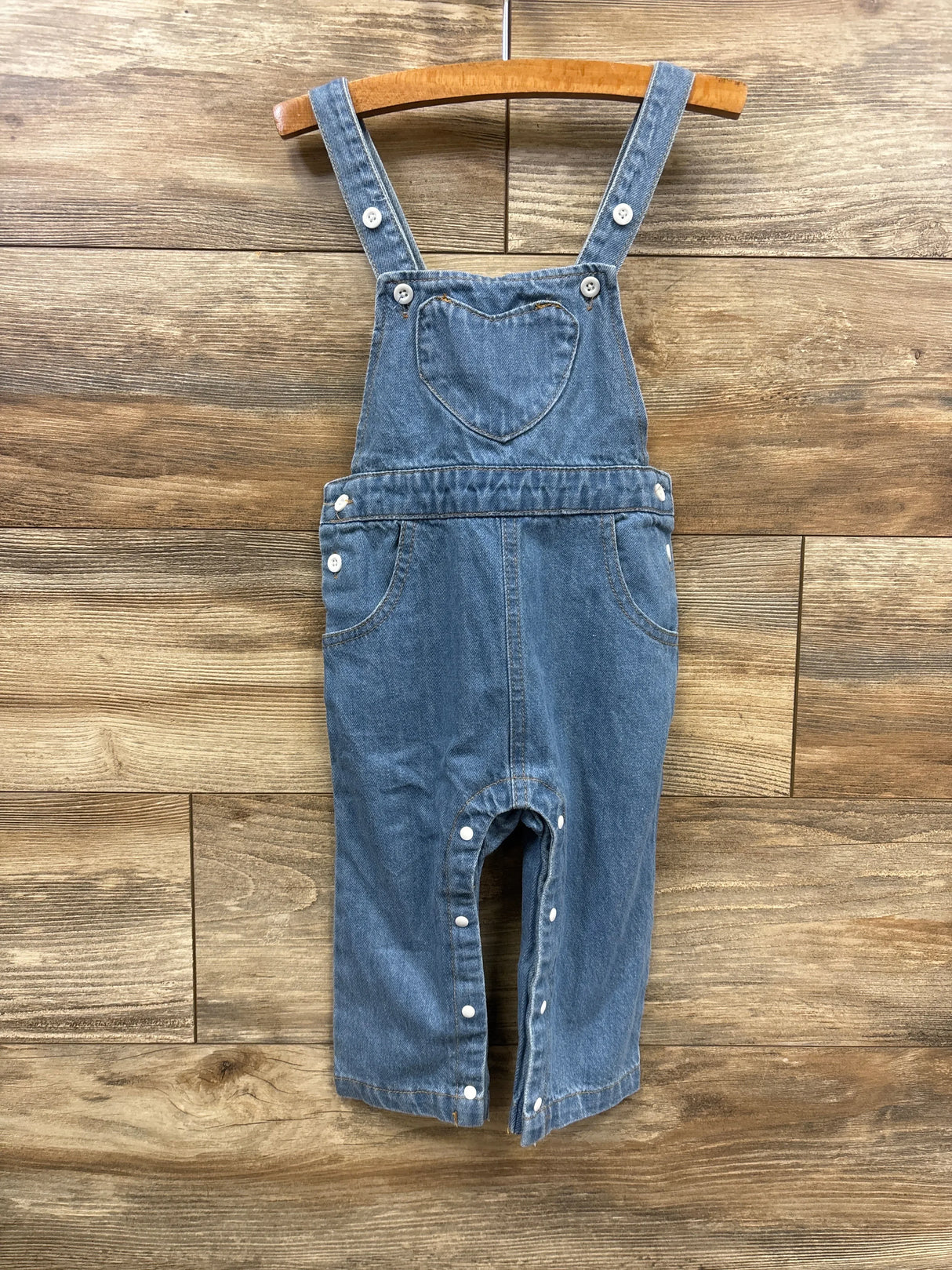 Blue denim toddler overalls with heart-shaped chest pocket and snap closures, hanging on wooden hanger
