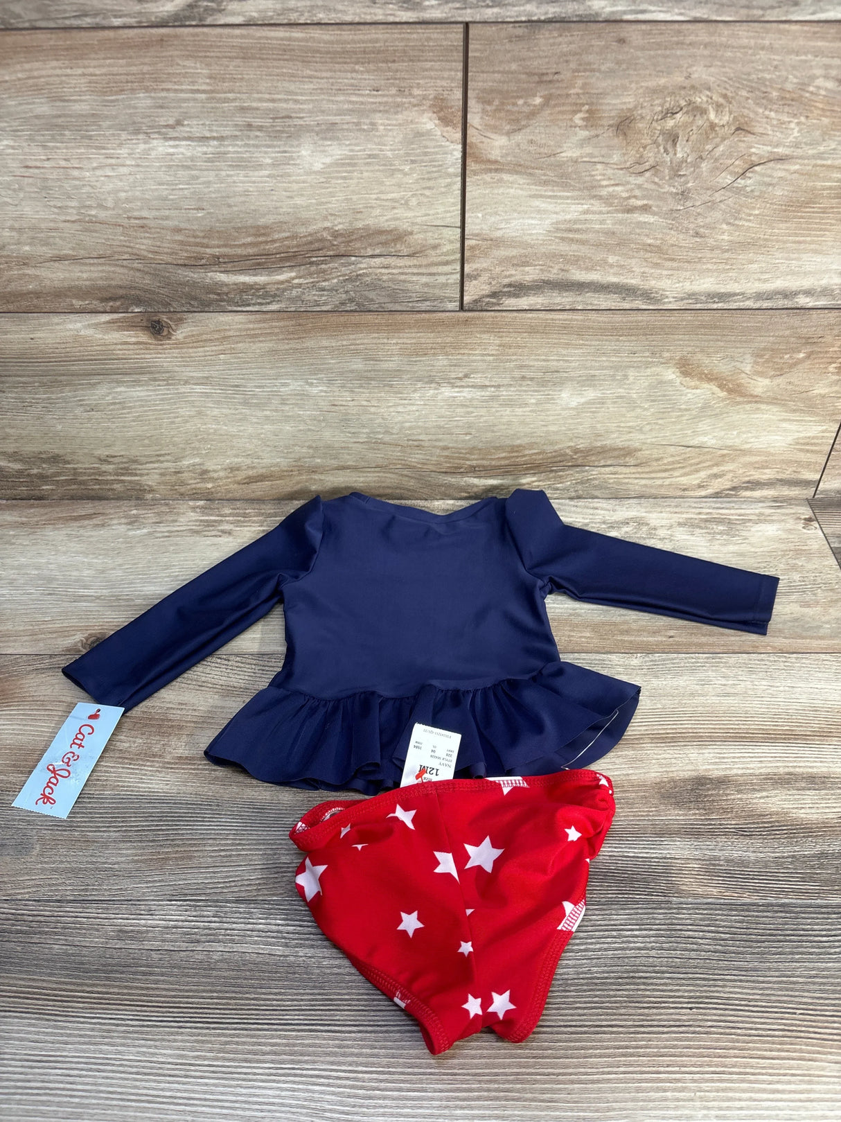 Flat lay of Cat & Jack 2-piece long-sleeve navy peplum rashguard with red star-print bottoms, size 12M on wood background