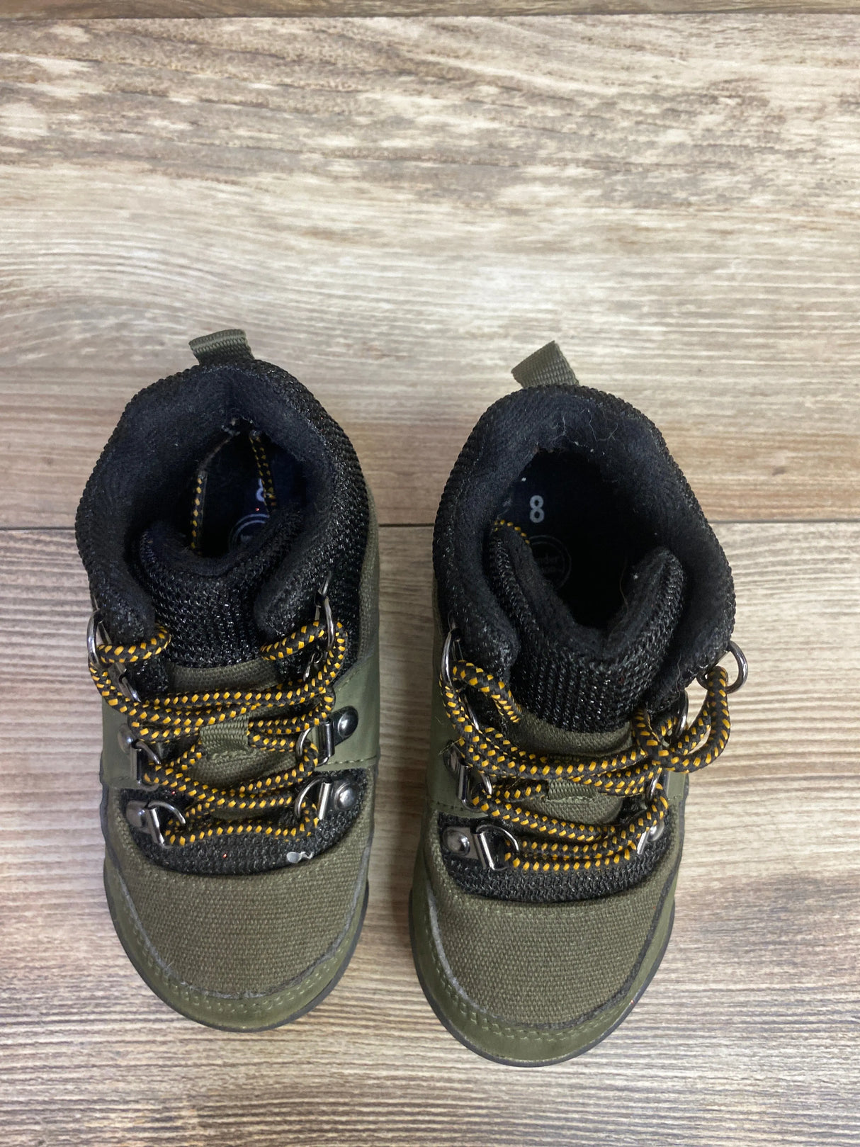 Pair of olive green toddler hiking boots with black padded collars and yellow-black laces on wood floor