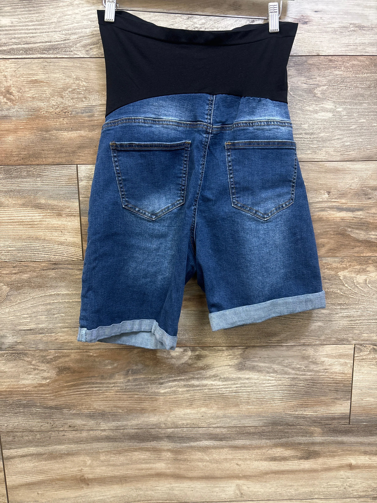 Back view of blue maternity denim shorts showcasing full black panel, back pockets, and cuffed hems on a wood floor