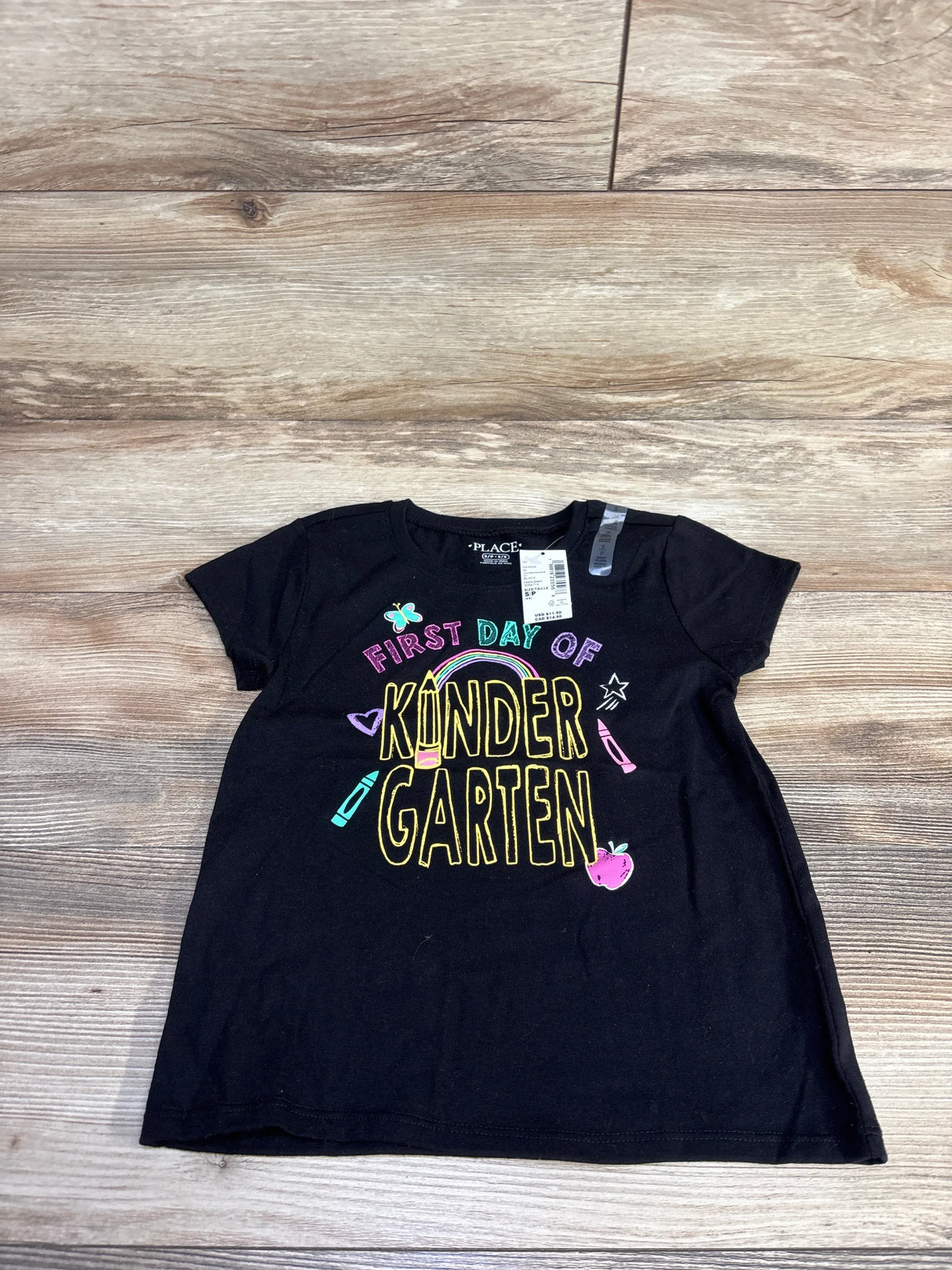 Flat lay of black Children's Place toddler tee with multicolored First Day of Kindergarten text and school icons on wood