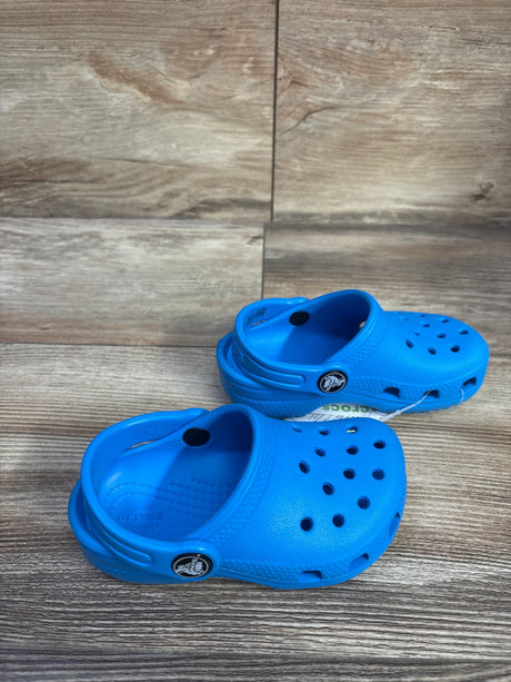 Front-left view of blue Crocs Classic clog size 7 showing heel strap and ventilation holes on wood background