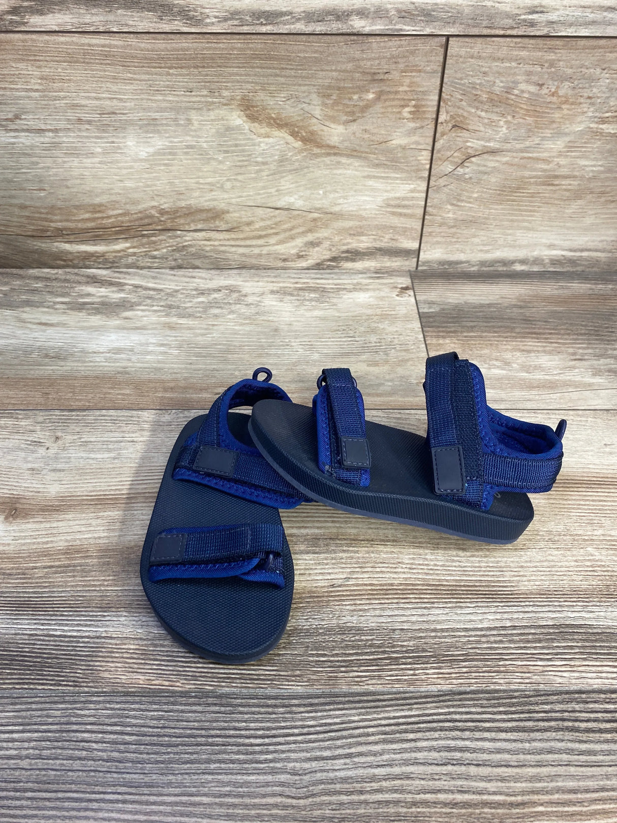 Angled front view of Old Navy toddler navy double-strap sandals showing adjustable hook-and-loop straps and textured footbed on wood floor