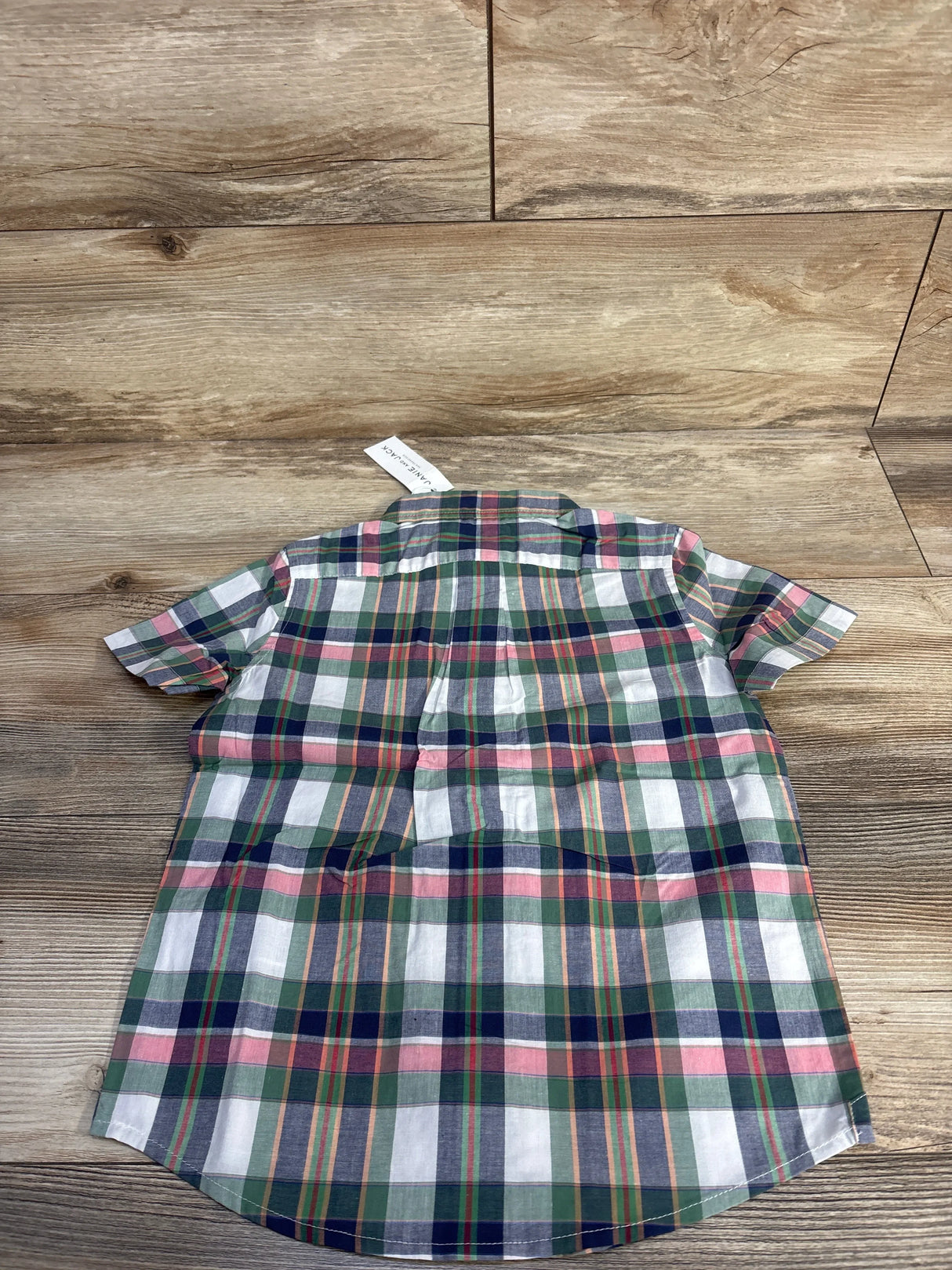 Back view of toddler short-sleeve Madras plaid button-down shirt in green showing box pleat and curved hem on wood background