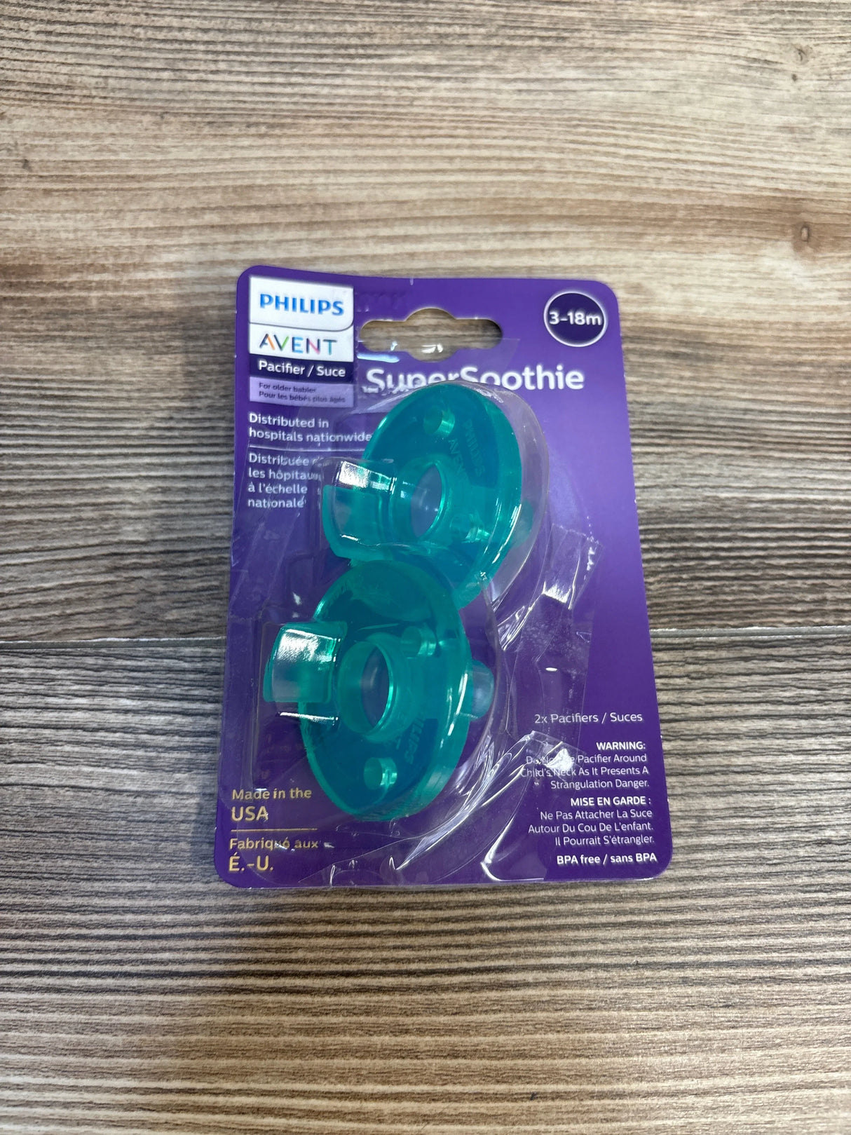 Front view of Philips Avent SuperSoothie 2pk pacifiers 3-18m showcasing teal silicone pacifiers in purple sealed blister card on wood plank floor