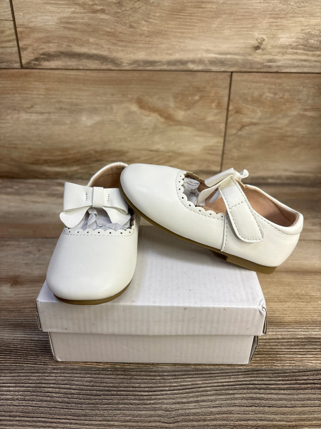 Front and side view of white leather baby princess flats showing scalloped trim and bow accent, on a white shoebox against a wood backdrop