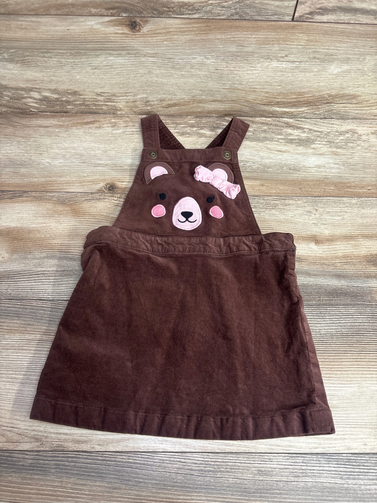 Brown toddler overall dress with pink bear face and ears embroidered on the front bib, laid on wood floor