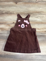 Brown toddler overall dress with pink bear face and ears embroidered on the front bib, laid on wood floor