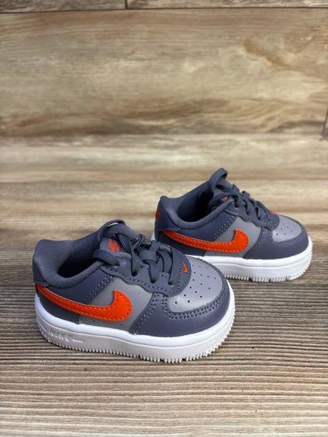 Pair of Nike Force 1 Low TD sneakers in cement grey and light carbon with orange Swooshes and white soles on wood flooring