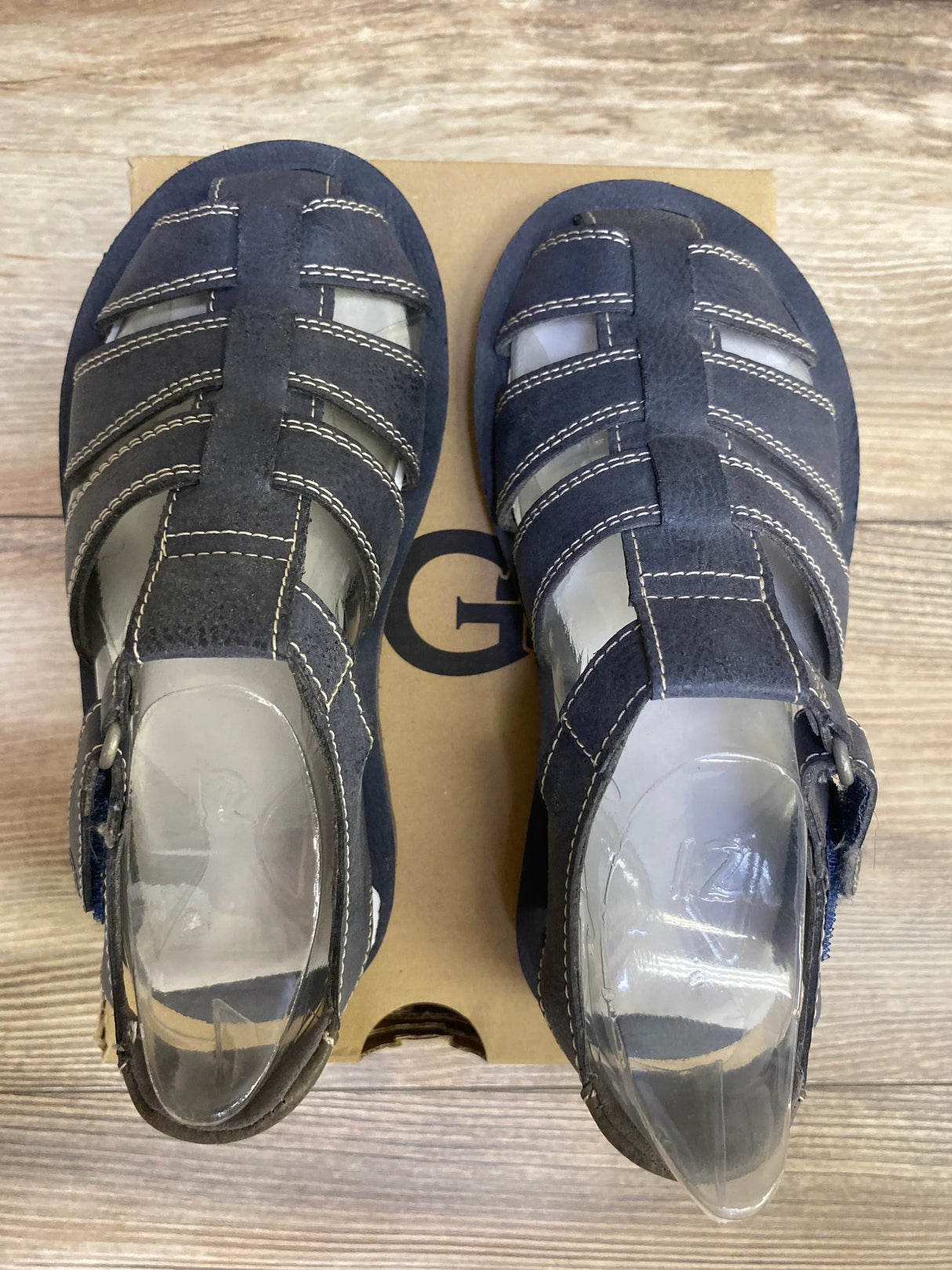 Top view of navy UGG Josiah fisherman toddler sandals with suede upper, white stitching and buckle straps on a shoebox