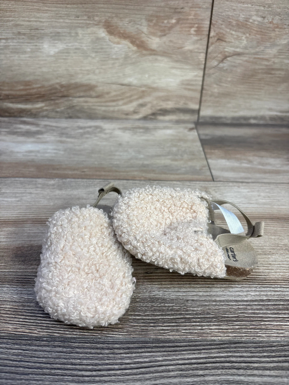 Angled top view of Child of Mine beige slip-on baby clog showing curly sherpa upper and elastic heel strap on wood-look background