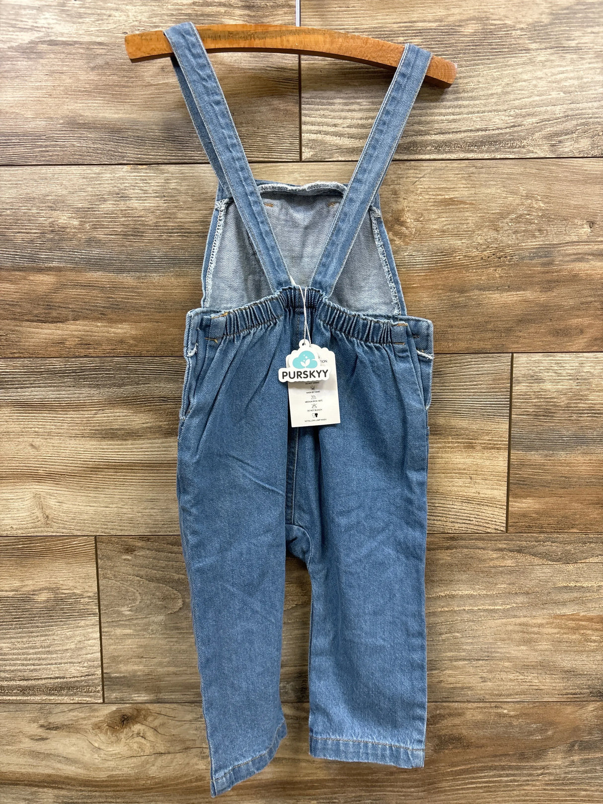 Kids blue denim overalls with adjustable straps hanging on wooden hanger