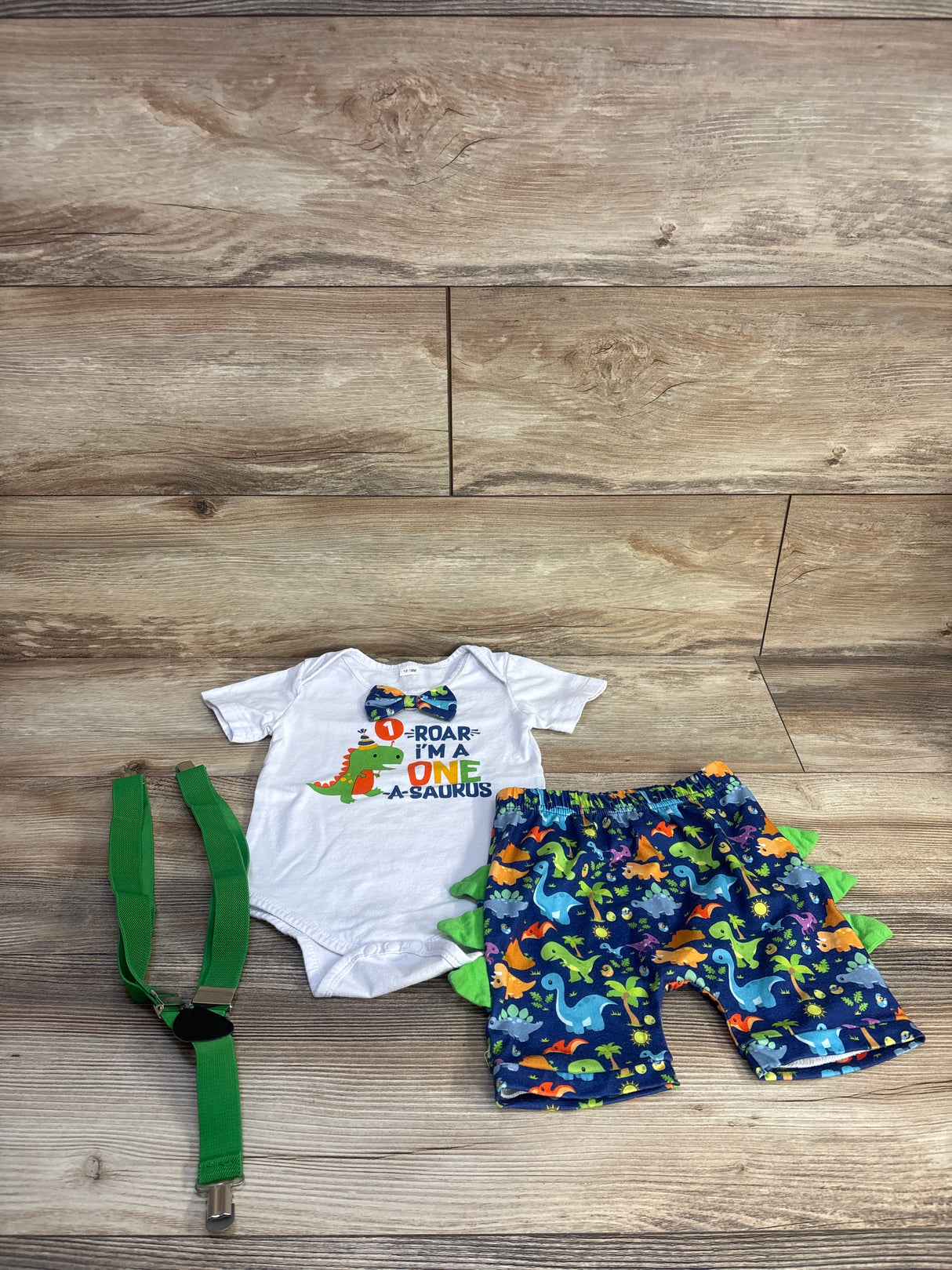 Flat lay of 3-piece baby 1st birthday suspender set: white onesie Roar I'm One, shorts with green spikes, green suspenders