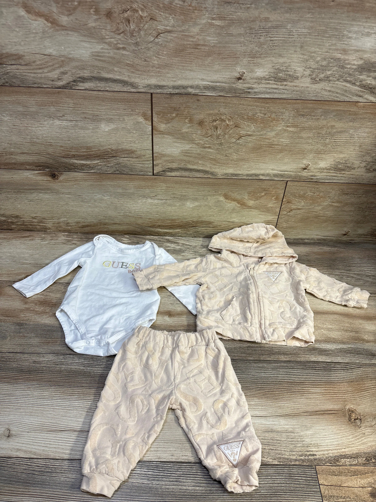 Front view of Guess Baby 3pc Zip-Up Jacket, Joggers, and Bodysuit Set in beige on a wood floor