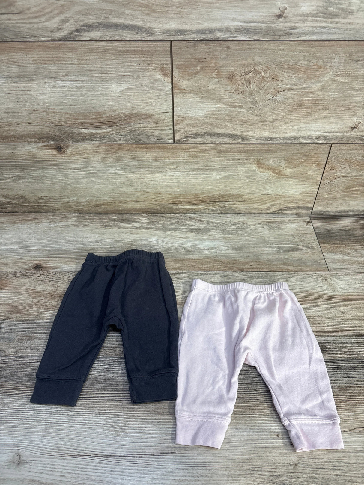 Top-down view of two Cloud Island newborn pull-on pants, grey and pale pink, showing elastic waist and cuffed ankles on wood background