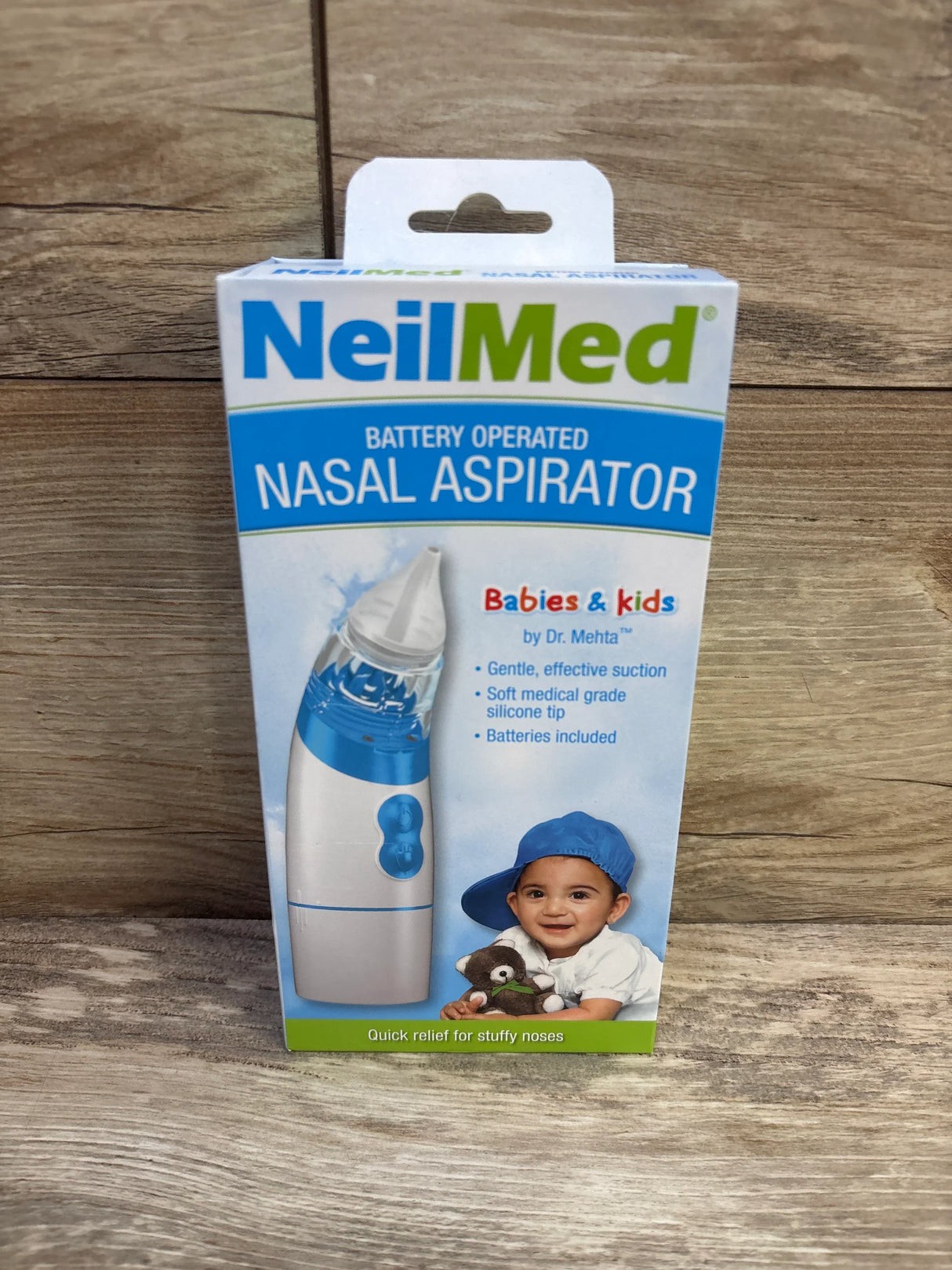 Front view of NeilMed battery operated nasal aspirator box showing the blue and white aspirator image and baby photo on a wooden background